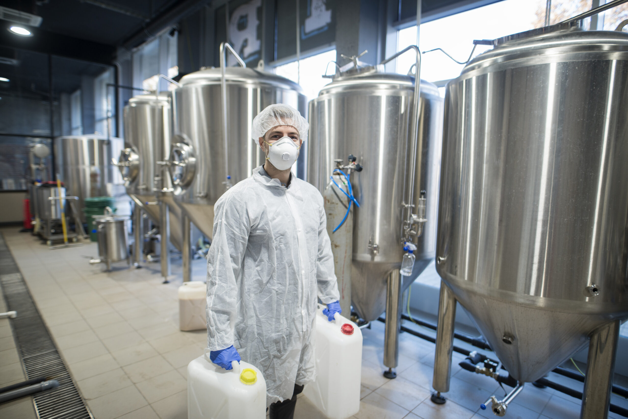 Industrial worker technologist in white protective suit with hairnet and mask holding plastic cans with chemicals in food factory production line.